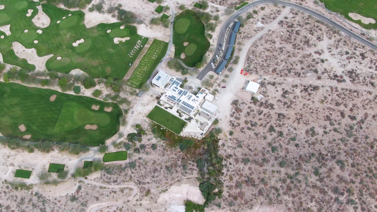 Drone view of a white structure beside golf greens and dry brush with a curved road above.