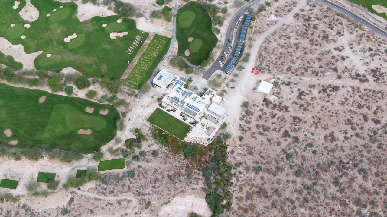 Mid-clip aerial frame over La Paz, Baja California Sur showing fairway shapes and bunkers along the upper and left edges, a white building near the road, dry brushland, and no visible people.