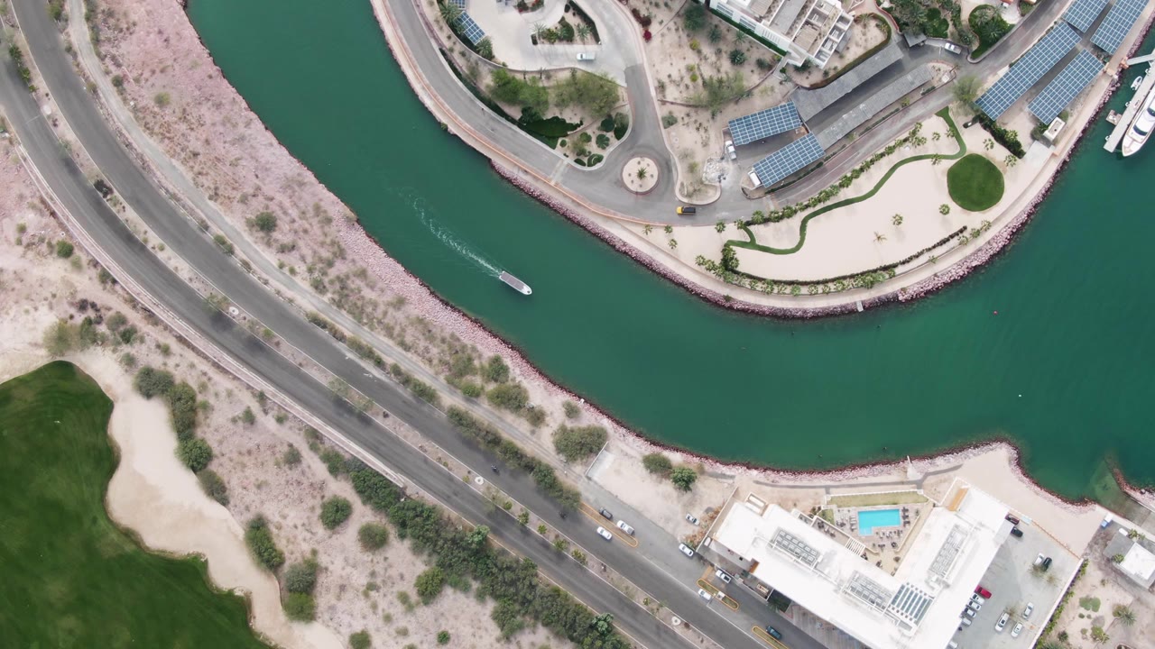 Overhead drone view in La Paz, Baja California Sur, showing a small boat entering a curved green canal beside a road, marina buildings, landscaped areas, desert ground, and no visible people.