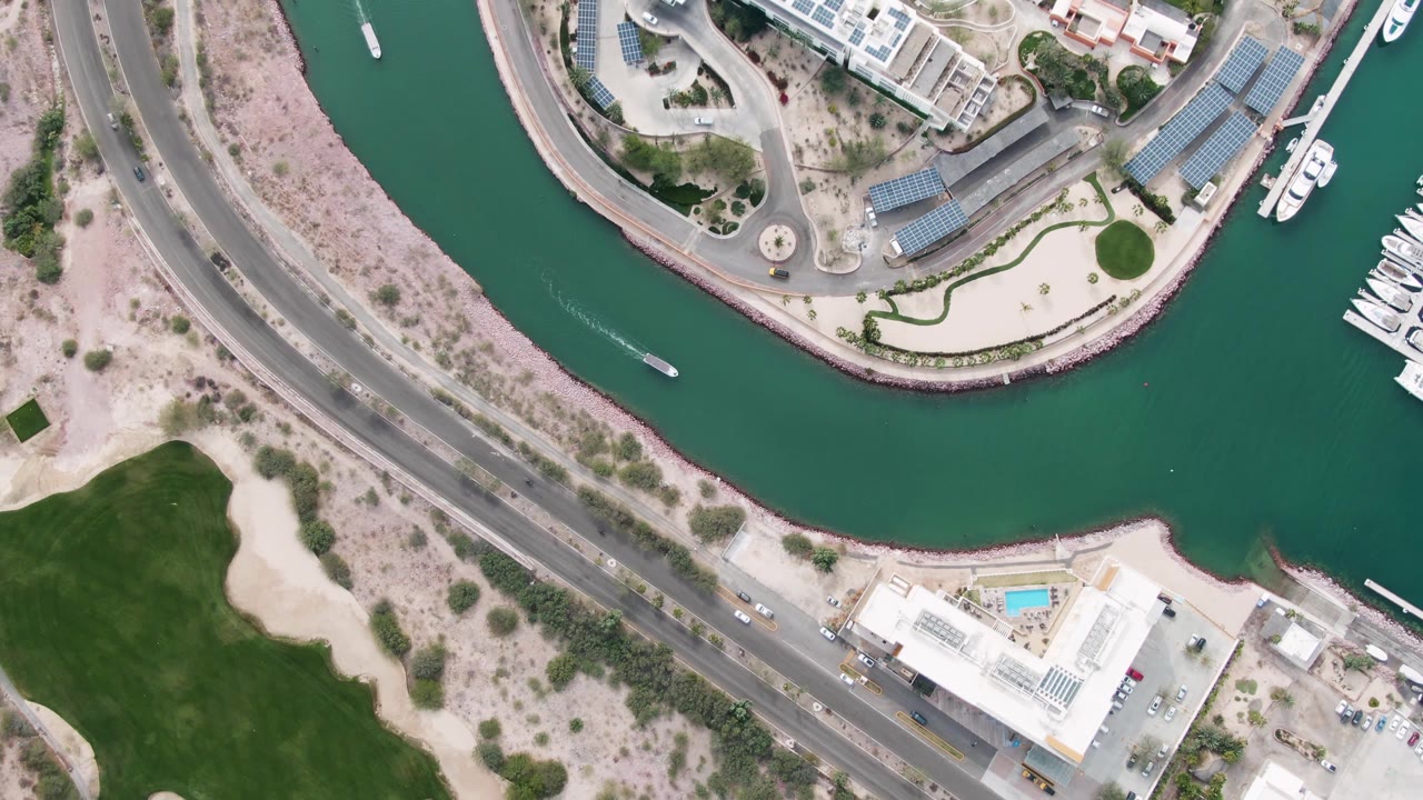 Top-down La Paz, Baja California Sur marina view with the boat continuing through the curved canal, docked yachts appearing at the right edge, road traffic below, pale ground, and no visible people.