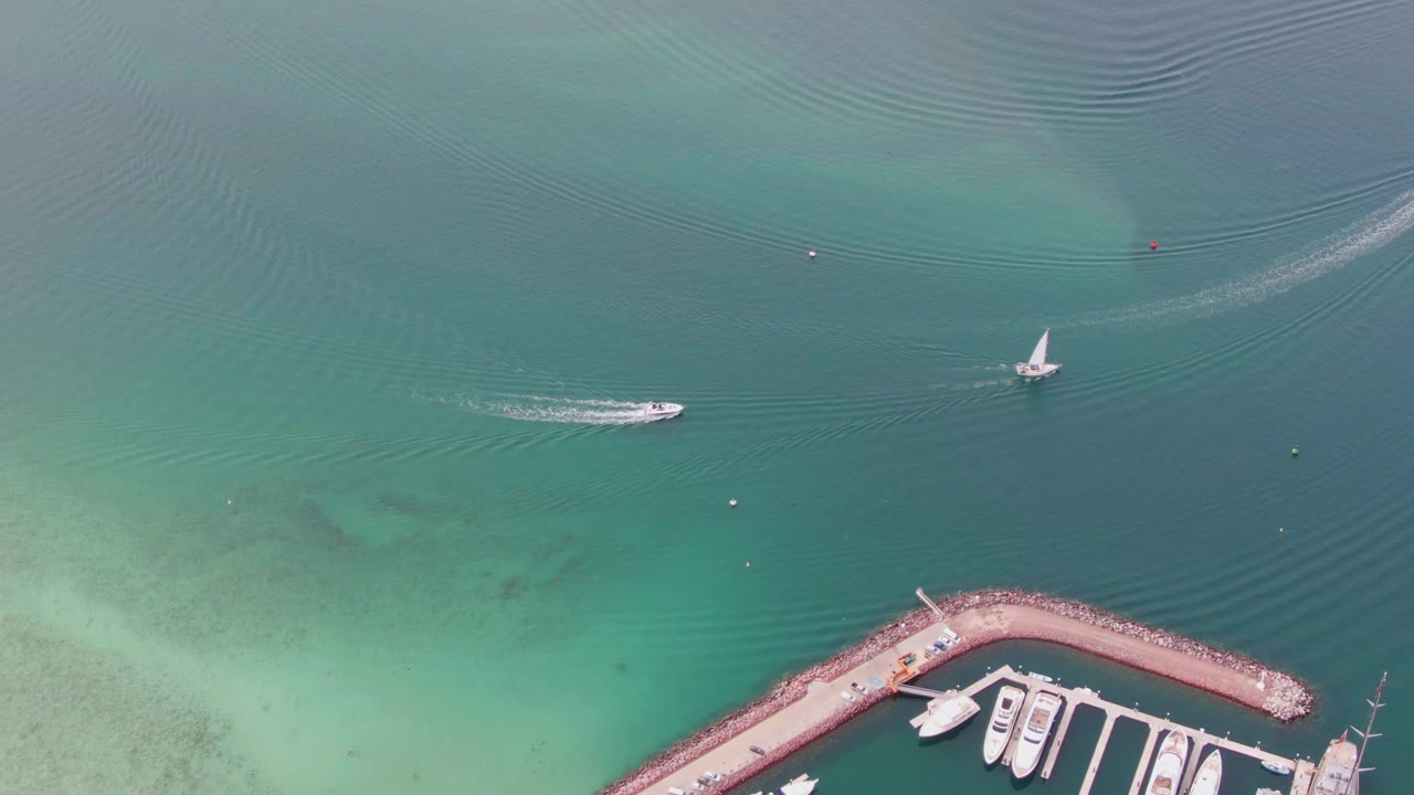 Aerial La Paz frame showing the speedboat slightly farther across the blue-green harbor water, the sailboat nearby, wake trails and ripple lines spreading across the surface, and no visible people.