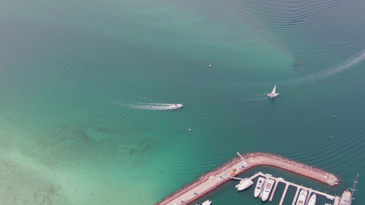 Overhead La Paz harbor view with the speedboat and its fading wake left of center, the sailboat farther right, soft green shallows below, marina docks in the lower corner, and no visible people.
