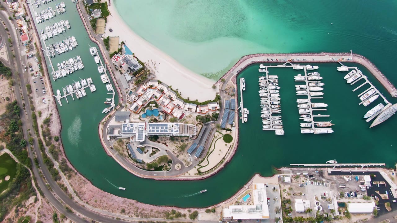 Top-down La Paz coastal marina view with yachts inside dark green water, a pale beach and turquoise water beyond the breakwater, road traffic at left, and no visible people.