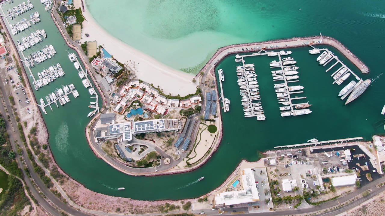 High aerial view of the La Paz marina with yacht rows, a sandy beach, turquoise shallows, a small boat on dark harbor water, and no visible people.