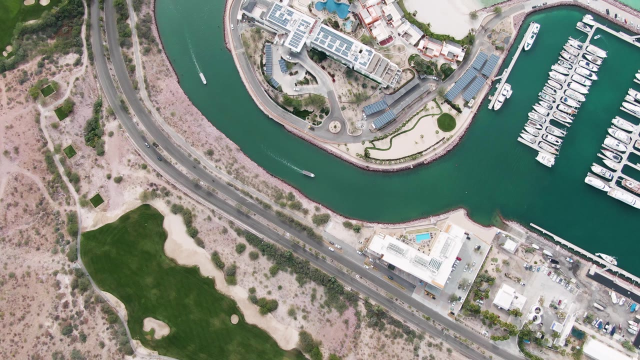 Overhead view in La Paz, Baja California Sur, showing a marina basin with docked white boats, a curved road, sandy terrain, green grass patches, buildings, and no visible people.
