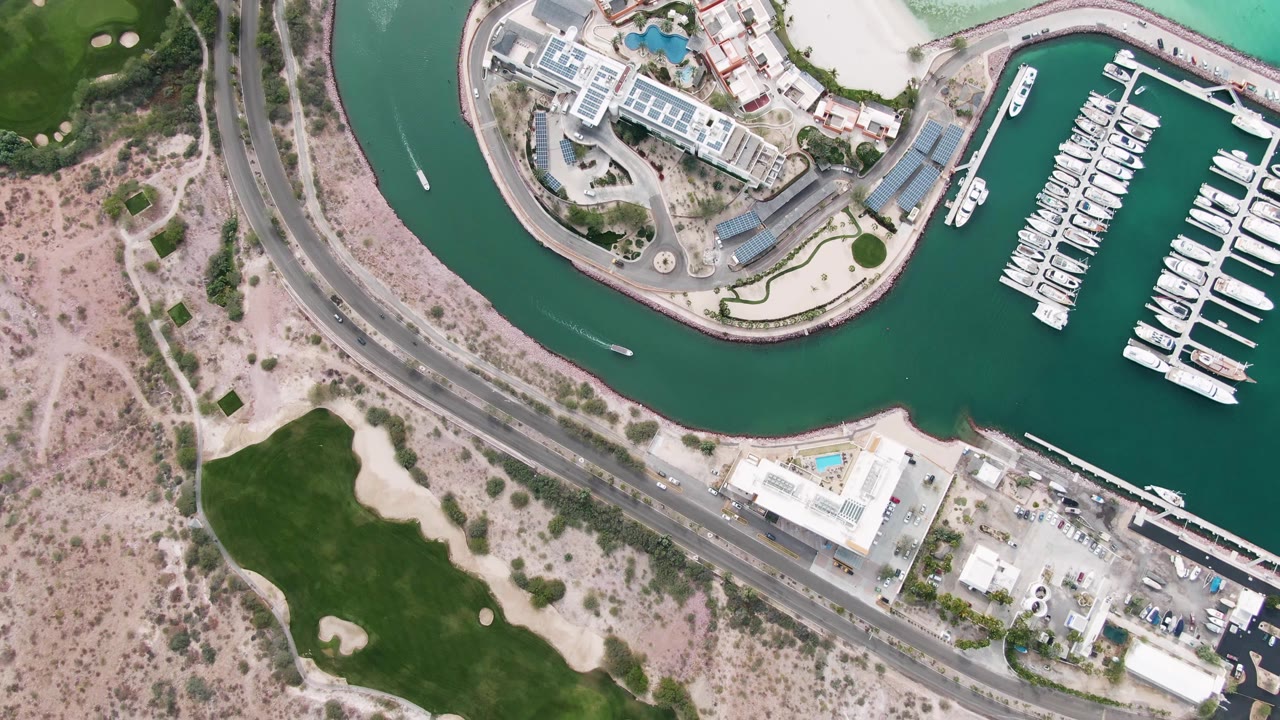 Aerial frame over La Paz with teal marina water on the right, docked boats in rows, a curved roadway through the middle, sandy terrain and grass on the left, and no visible people.