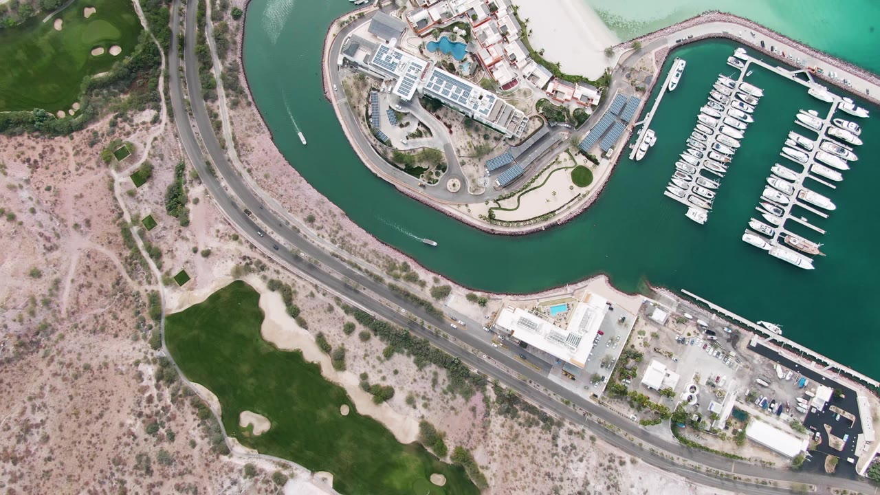 Top-down La Paz marina view with a small moving boat leaving a white wake in the channel, docked boats to the right, a curved road below, sandy terrain, green grass, and no visible people.