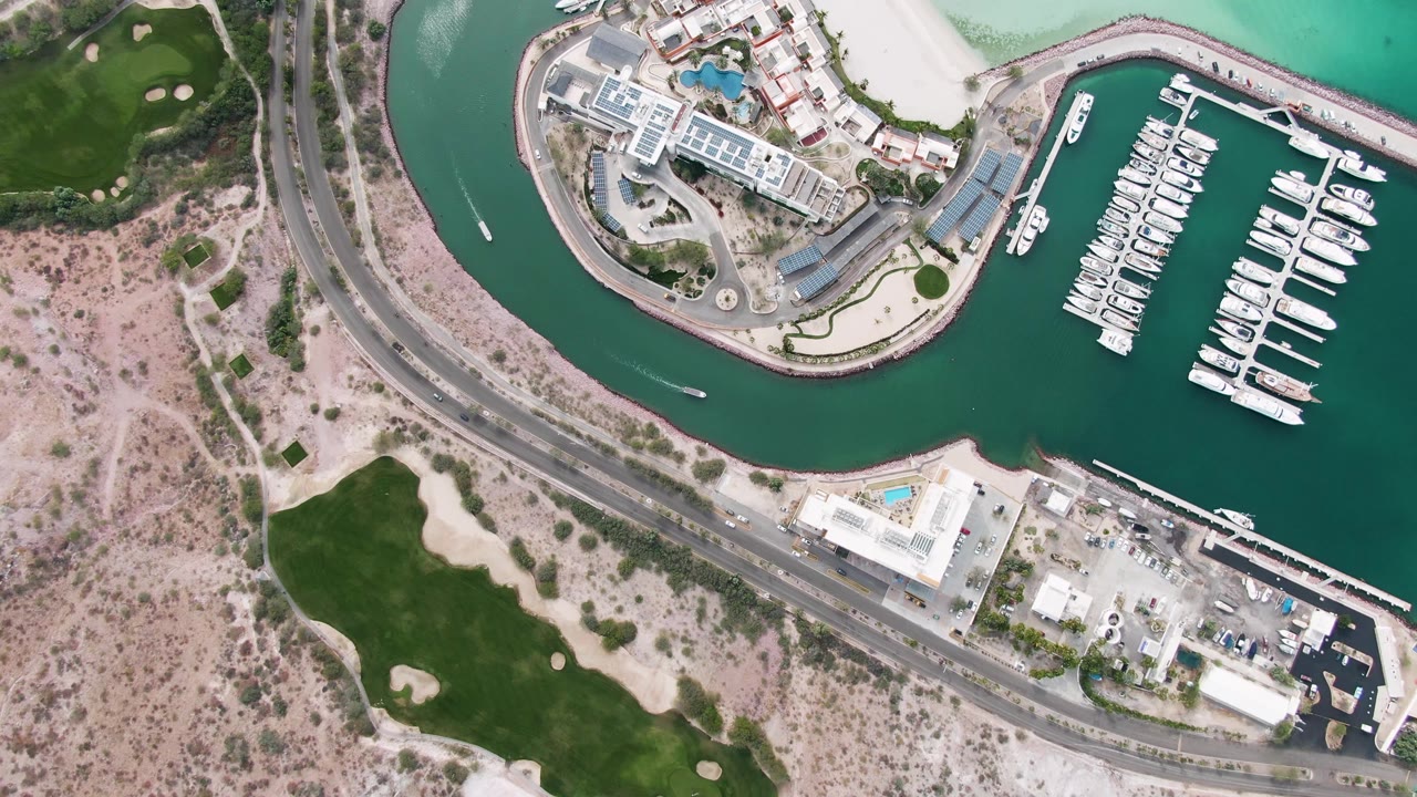 Overhead view of marina water, boat docks, coastal road, sandy terrain, and grass.