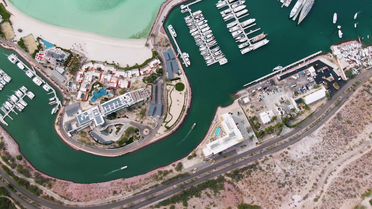 Aerial frame over La Paz showing white boats docked in dark green marina water, turquoise shallows at the beach edge, waterfront structures, a road, dry land, and no visible people.