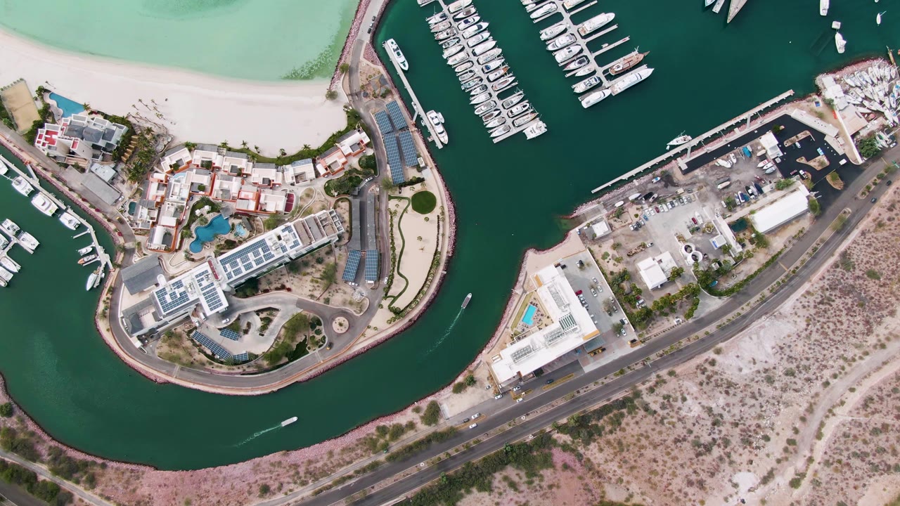 Marina channel, docked boats, beachfront buildings, road, and pale sand from above.