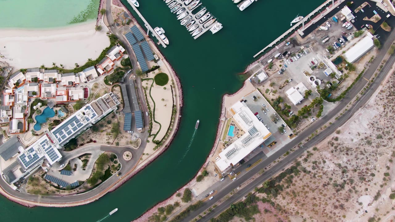 Overhead view in La Paz, Baja California Sur, showing a canal between waterfront buildings, marina docks, a road, dry land, and a small boat wake near the left side with no visible people.