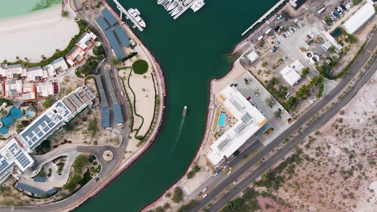 Overhead La Paz canal view with the boat wake now lower in the waterway, marina docks near the top edge, waterfront buildings with solar panels, a paved road, and scrubby dry terrain with no visible people.