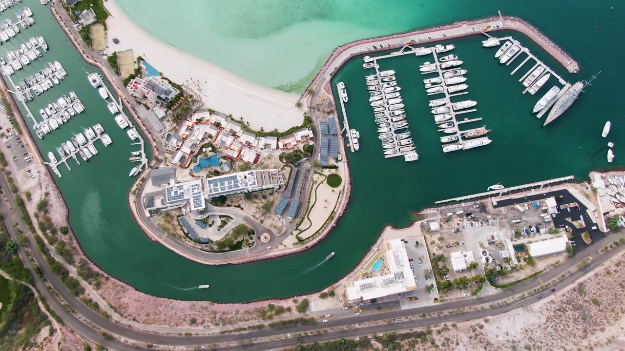 Second-frame aerial view over La Paz marina docks, with rows of moored boats, darker harbor water, pale beach water above, roadside traffic below, and no visible people.