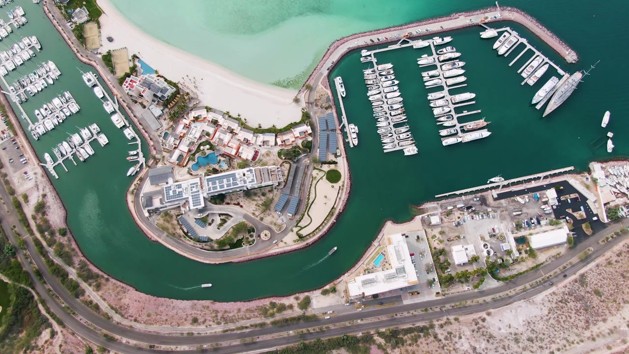 Top-down La Paz harbor frame showing the curved protective wall, packed marina slips, nearby beach, waterfront buildings, coastal roadway, and no visible people.