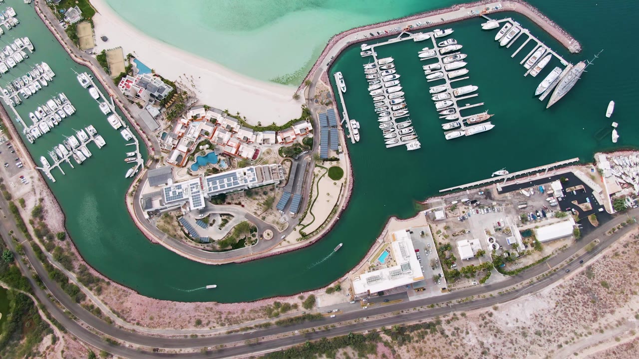 Mid-clip overhead view of the La Paz marina area with docked boats, a blue pool-like courtyard feature, waterfront buildings, road traffic, and no visible people.