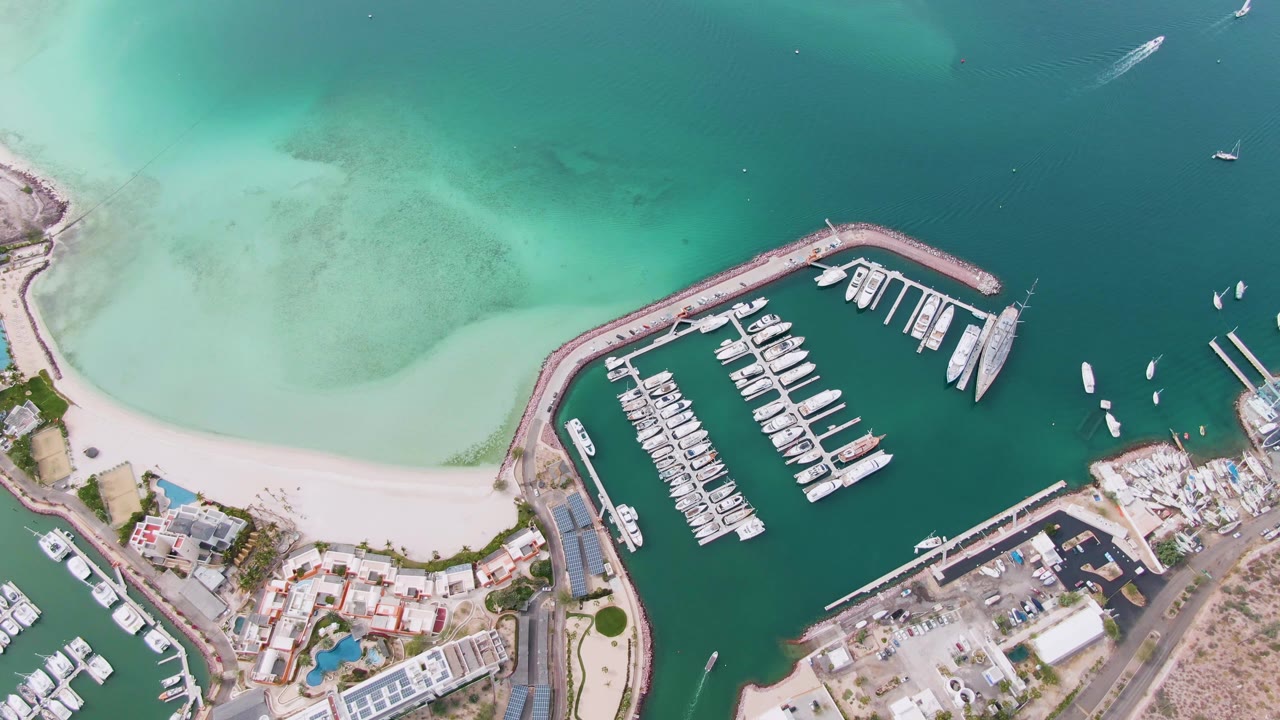 Overhead view in La Paz, Baja California Sur, showing a marina packed with docked boats, a curved breakwater, pale beach, turquoise shallows, shoreline buildings, and no visible people.