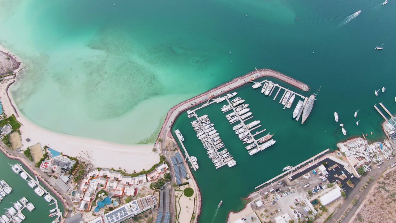 Aerial marina basin with rows of boats, sandy beach, and shallow turquoise water.