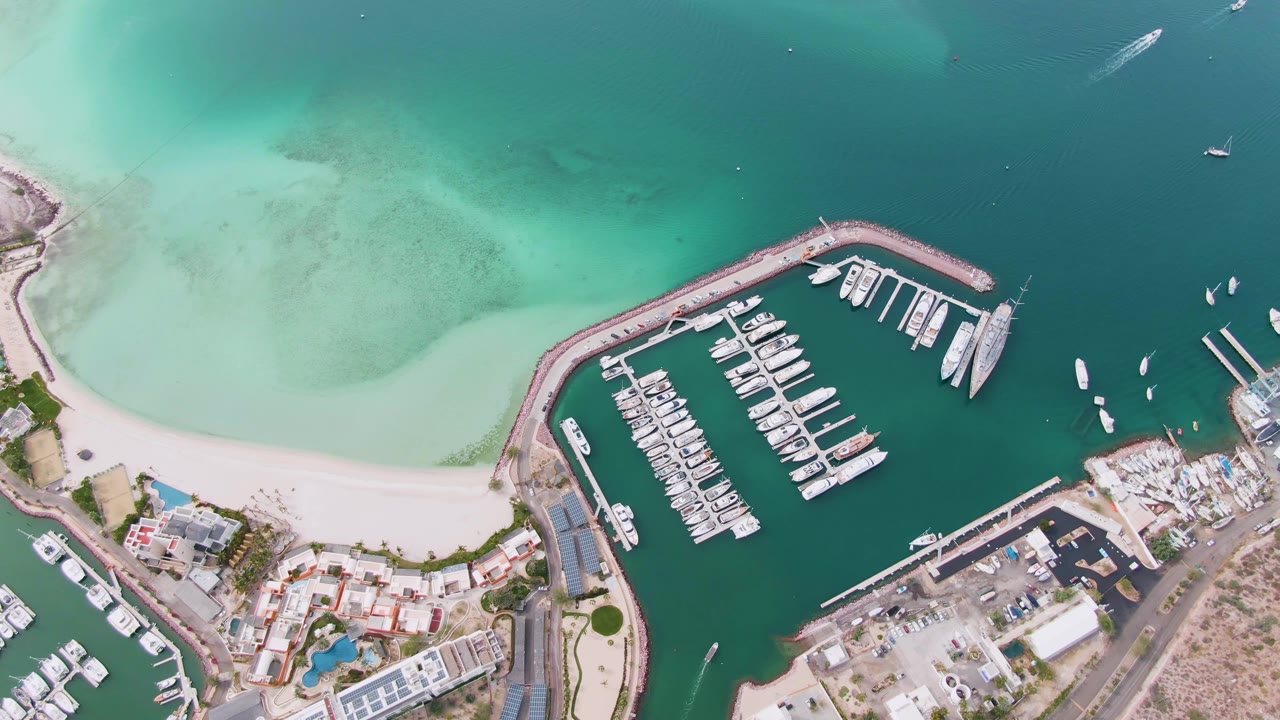 Drone-style overhead view in La Paz with the curved marina breakwater arcing through frame, clustered white boats in slips, open green-blue water to the right, pale beach to the left, and no visible people.