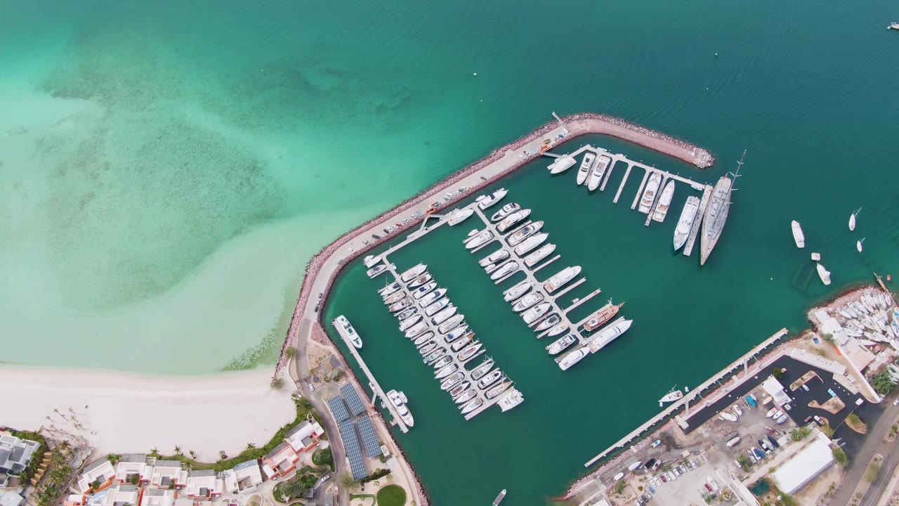 Top-down La Paz marina view with the breakwater arcing across the frame, rows of white boats, pale shoreline sand, mottled turquoise water currents, and no visible people.