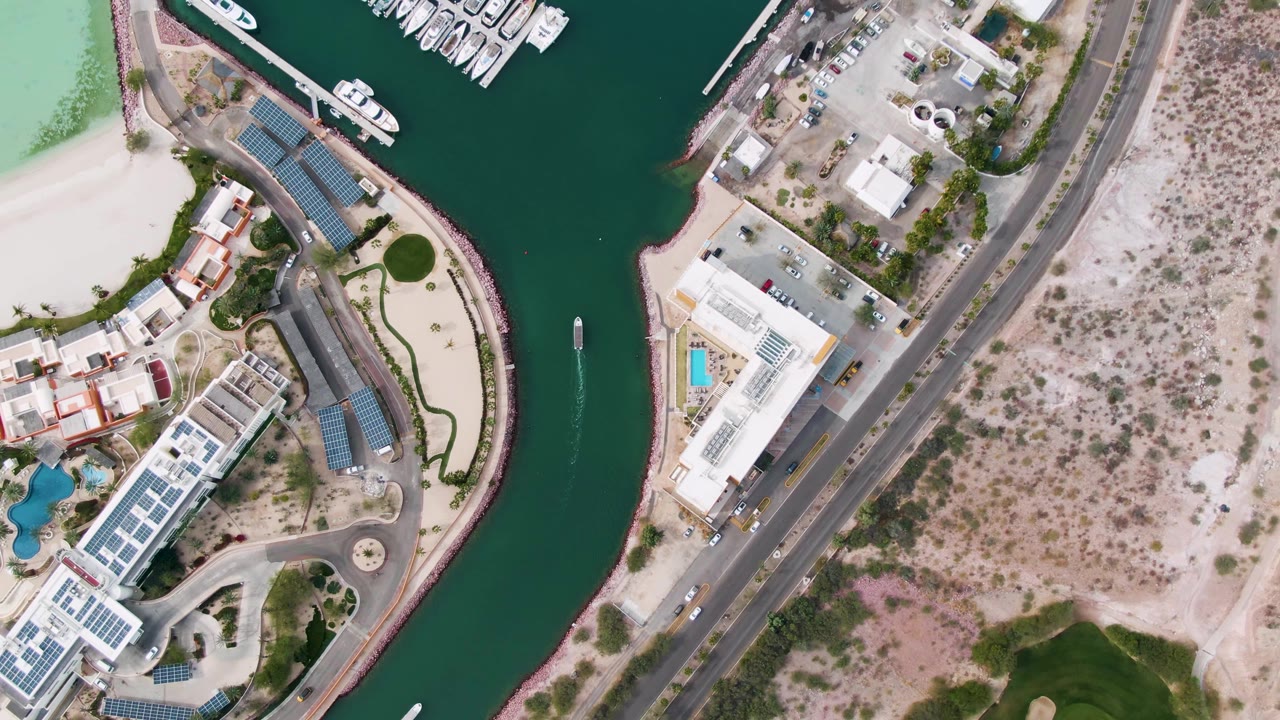 Overhead La Paz marina view with docked boats near the top edge, pale-roofed buildings and pools beside the canal, a paved road, scrubby desert ground, and no visible people.