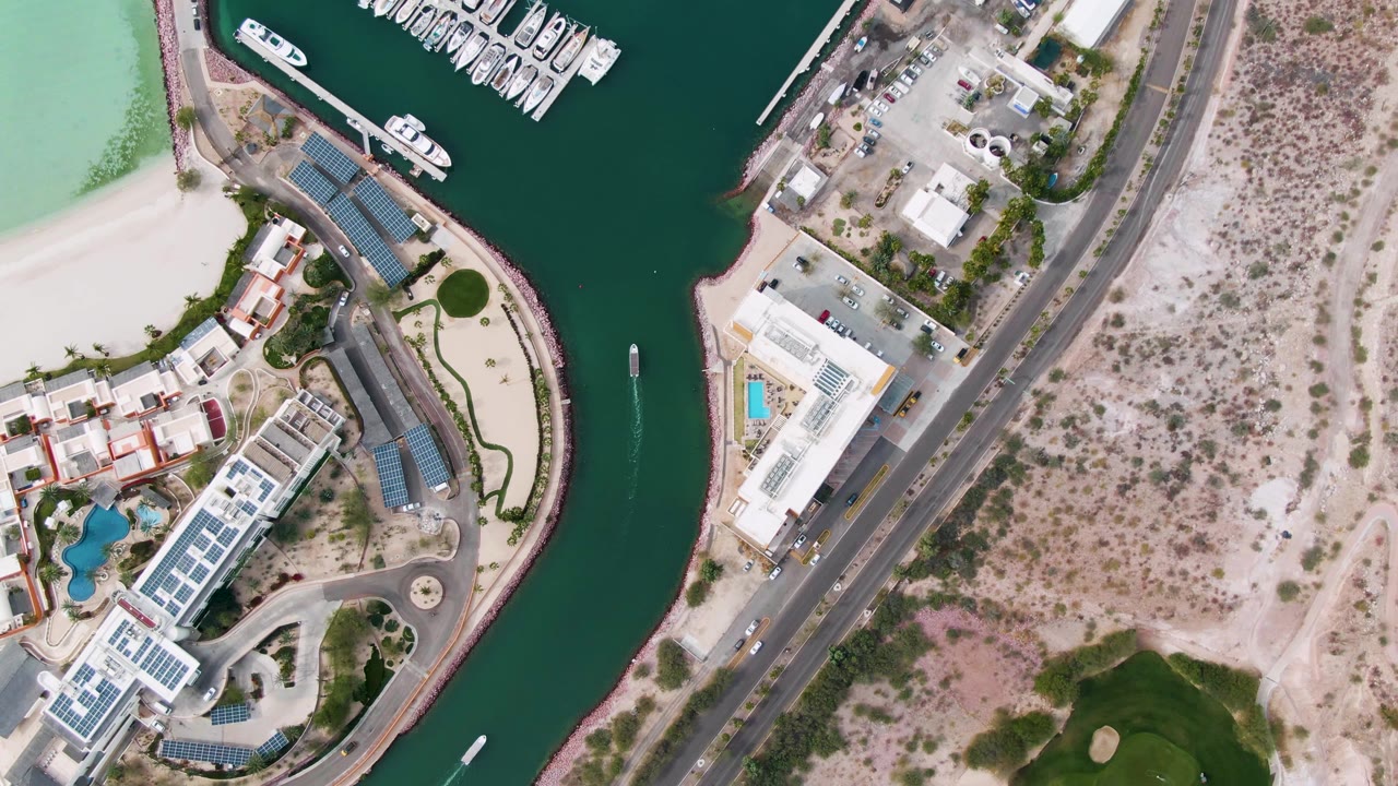Aerial frame over La Paz showing the marina docks at the upper edge, green canal water, waterfront buildings with pool areas, a road through sandy terrain, and no visible people.
