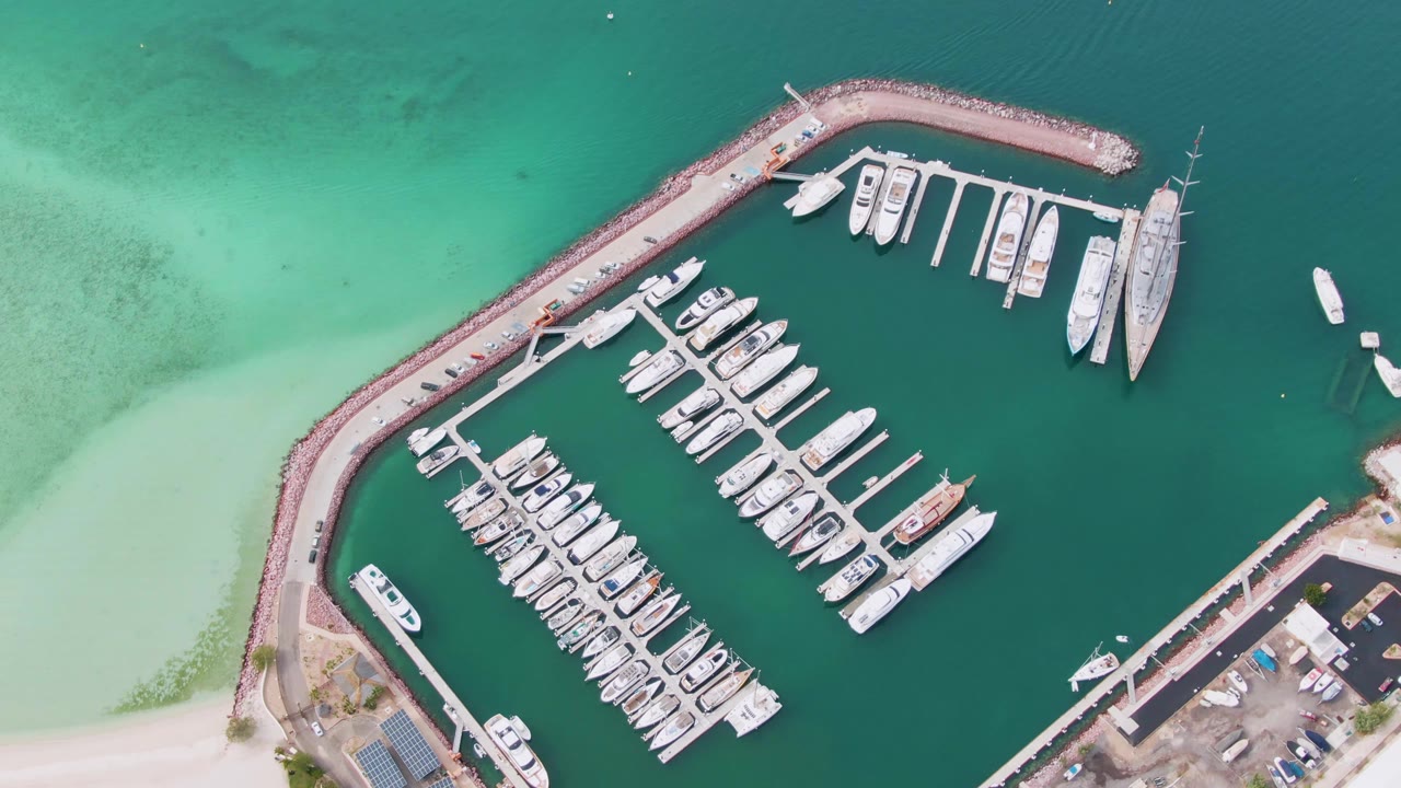 Overhead view of a La Paz marina with rows of docked white boats, a curved breakwater, shallow turquoise water at left, shore facilities at right, and no visible people.