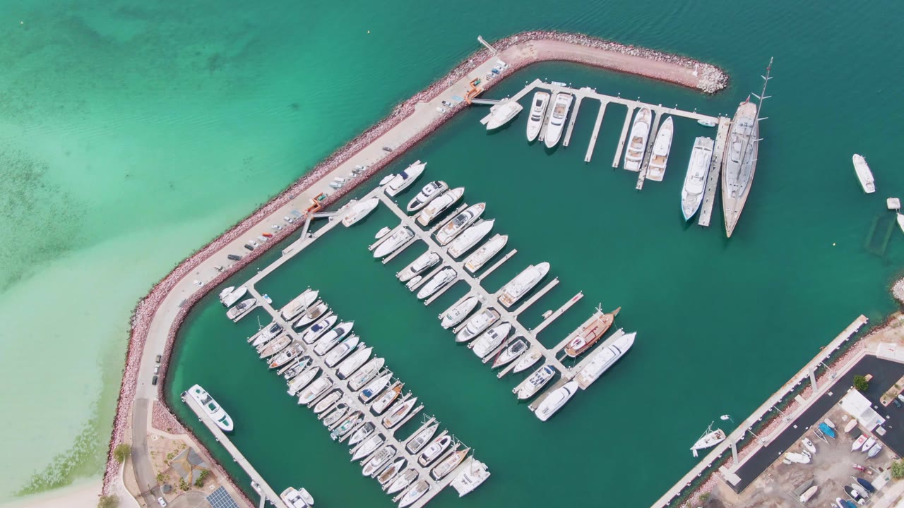 A La Paz marina is seen from above with the curved harbor wall enclosing docked boats, darker green water in the basin, pale shallow water near the beach edge, and no visible people.