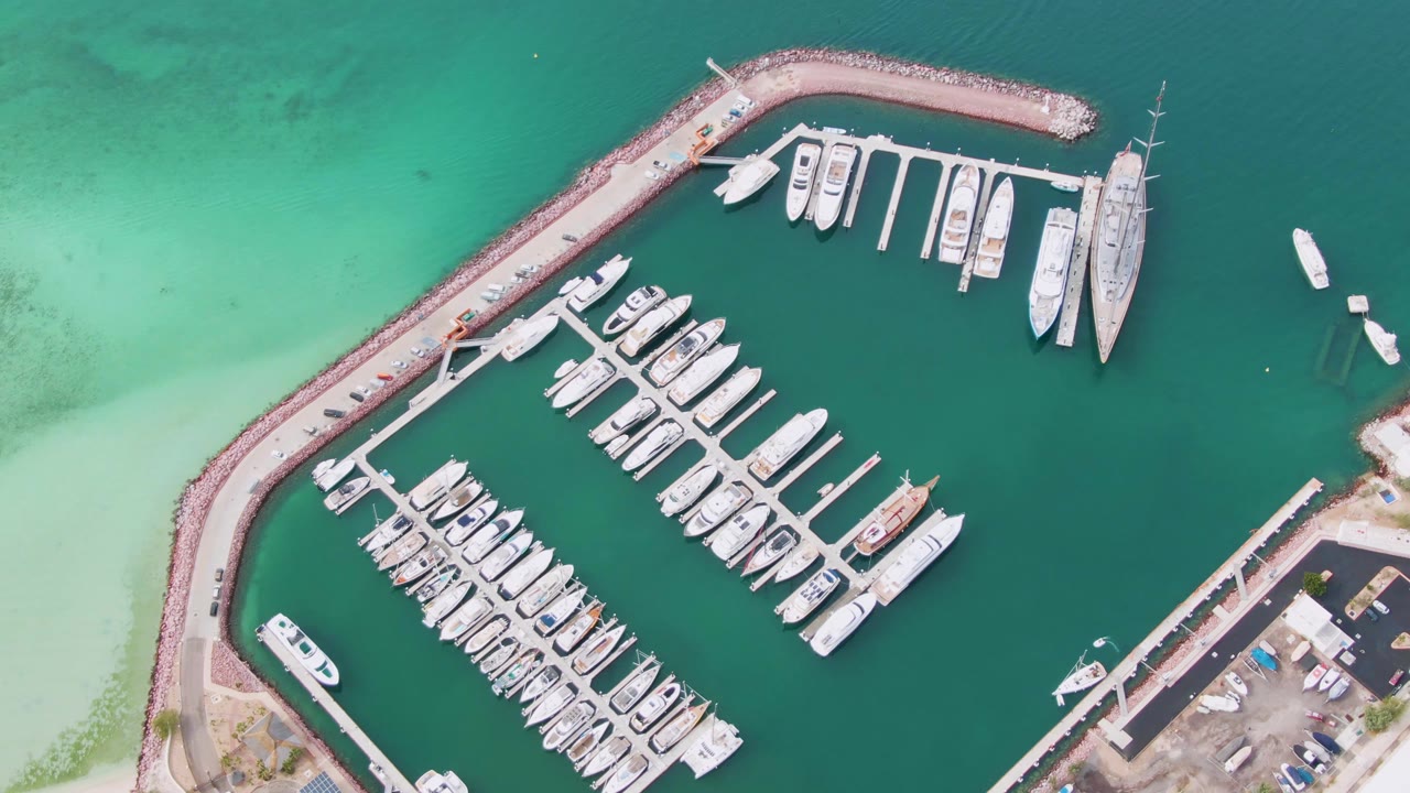 Overhead frame of the La Paz marina with docked boats arranged in rows, a small boat near the open water at upper right, shore structures at lower right, and no visible people.