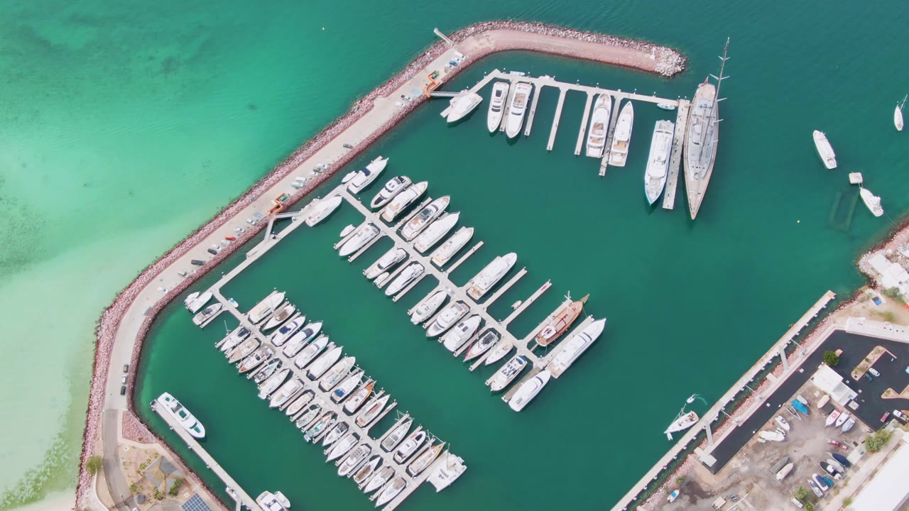 Top-down view of the La Paz marina with the curved breakwater sweeping around rows of white boats, pale water along the left shoreline, parked cars on the right shore, and no visible people.