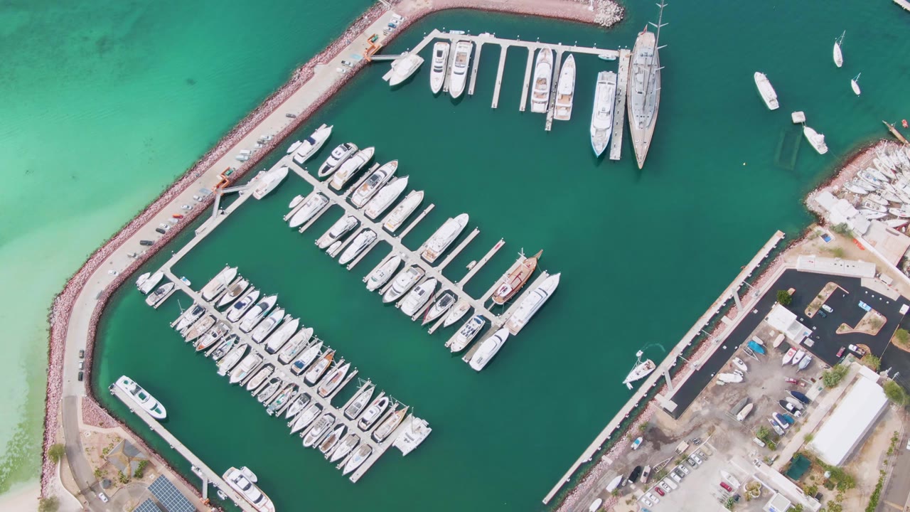 Overhead view of a La Paz, Baja California Sur marina at the start of the clip, with rows of docked boats in green water, a curved breakwater, shoreline roads, parking areas, and no visible people.