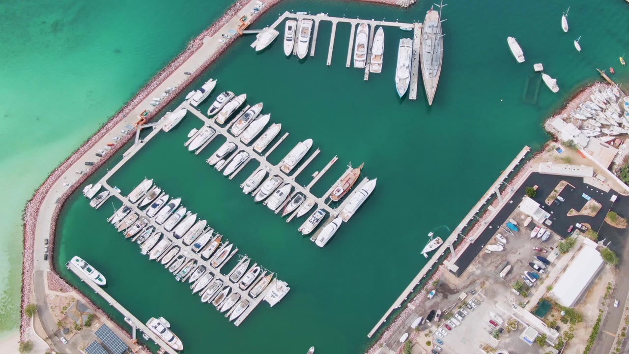 Overhead La Paz marina frame with tightly packed docked boats, open green water to the right, shoreline parking and service areas along the lower edge, and no visible people.