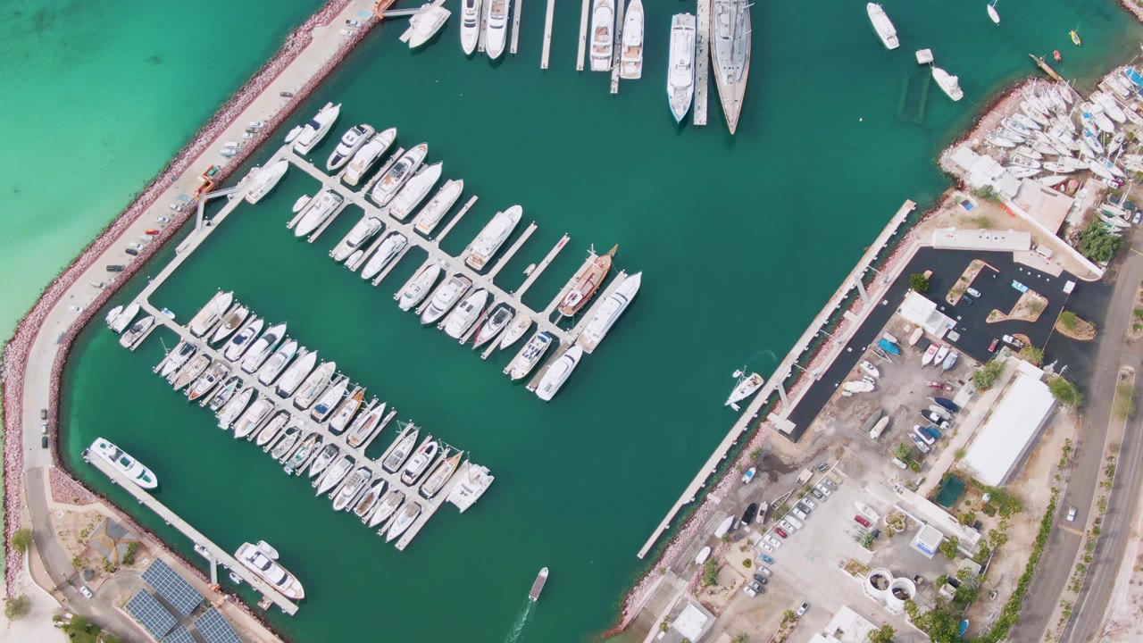 Overhead view of marina docks, moored boats, breakwater, roads, and green coastal water.