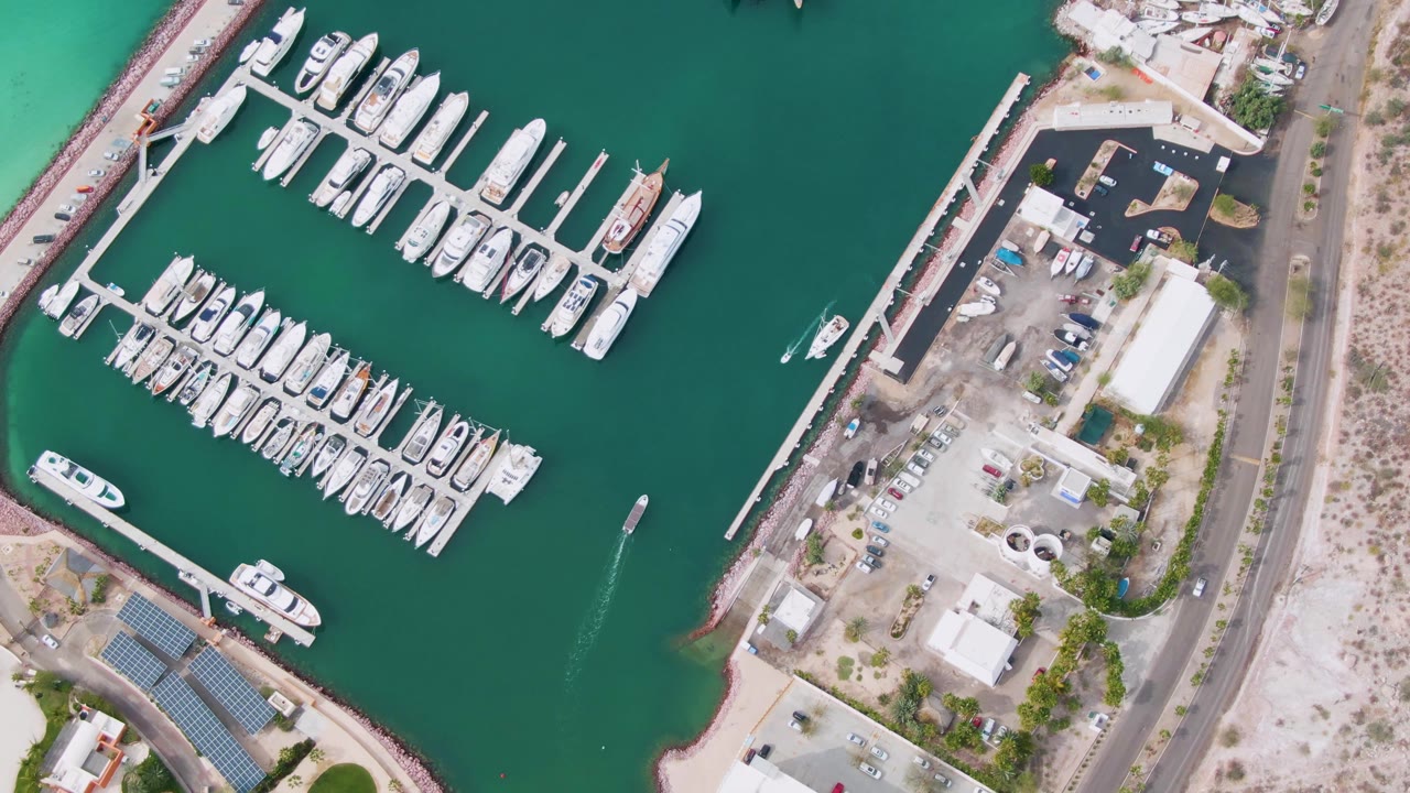 Overhead view of a La Paz marina with white yachts docked in rows, green water, shoreline roads, parking areas, solar-panel roofs, and no visible people at the start of the clip.