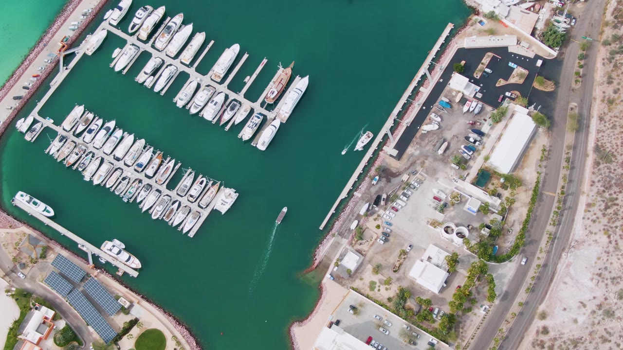 Aerial marina view with rows of yachts, a small boat wake, parking, and shoreline road.
