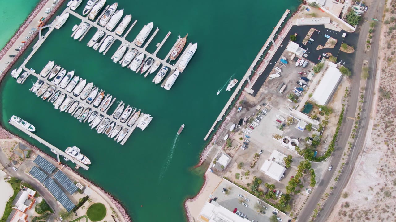 Top-down La Paz marina frame with white yachts aligned along piers, a long dock edge, green water, shoreline service areas, roads, parking spaces, and no visible people.