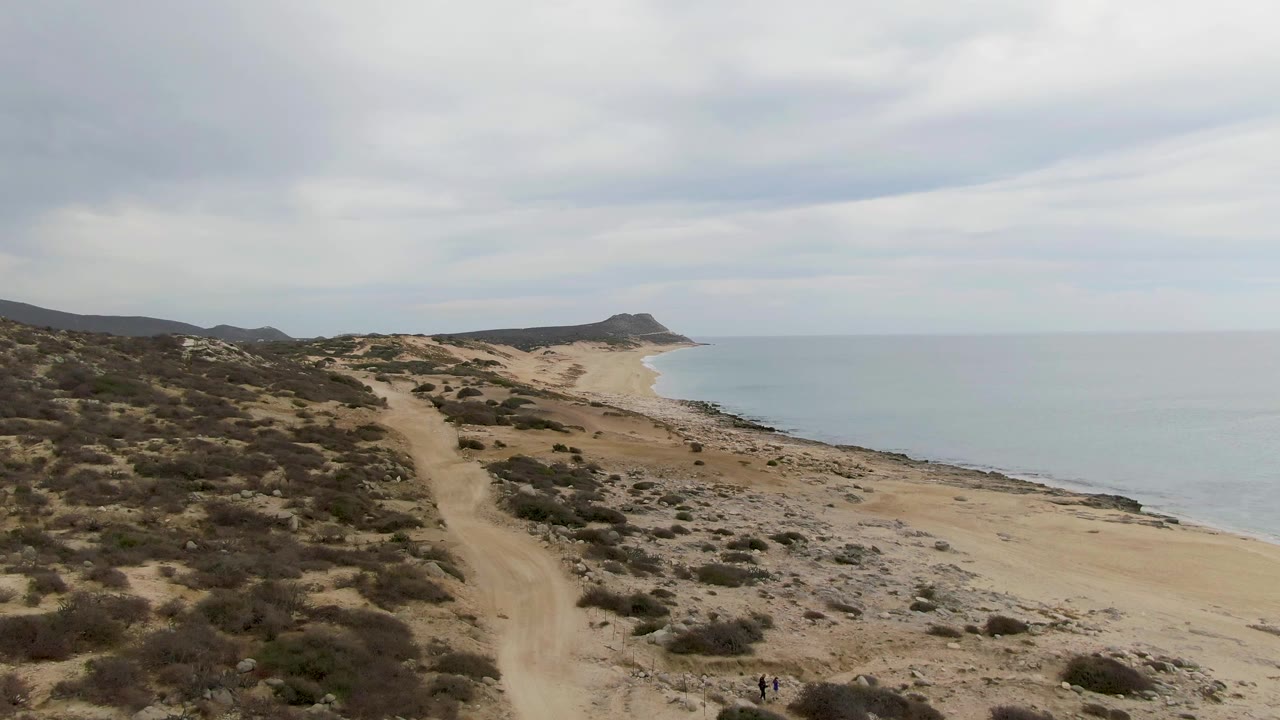 Elevated Punta Gorda Beach view with scrubby dunes dominating the left side, a pale sandy shoreline and blue-gray ocean on the right, distant hills on the horizon, and tiny people near the lower edge.