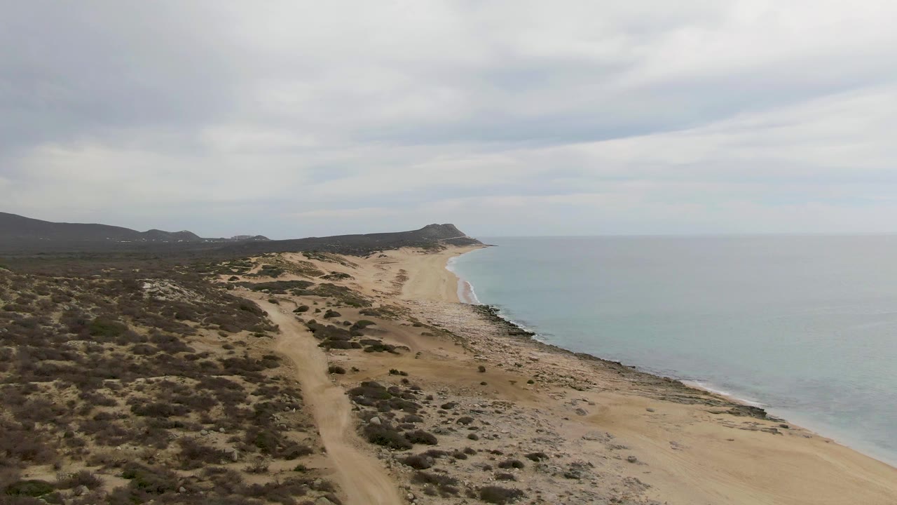 Opening aerial view over Punta Gorda Beach, Baja California Sur, with scrubby dunes and a dirt path leading toward a sandy shoreline, pale ocean, distant hills, and no visible people.