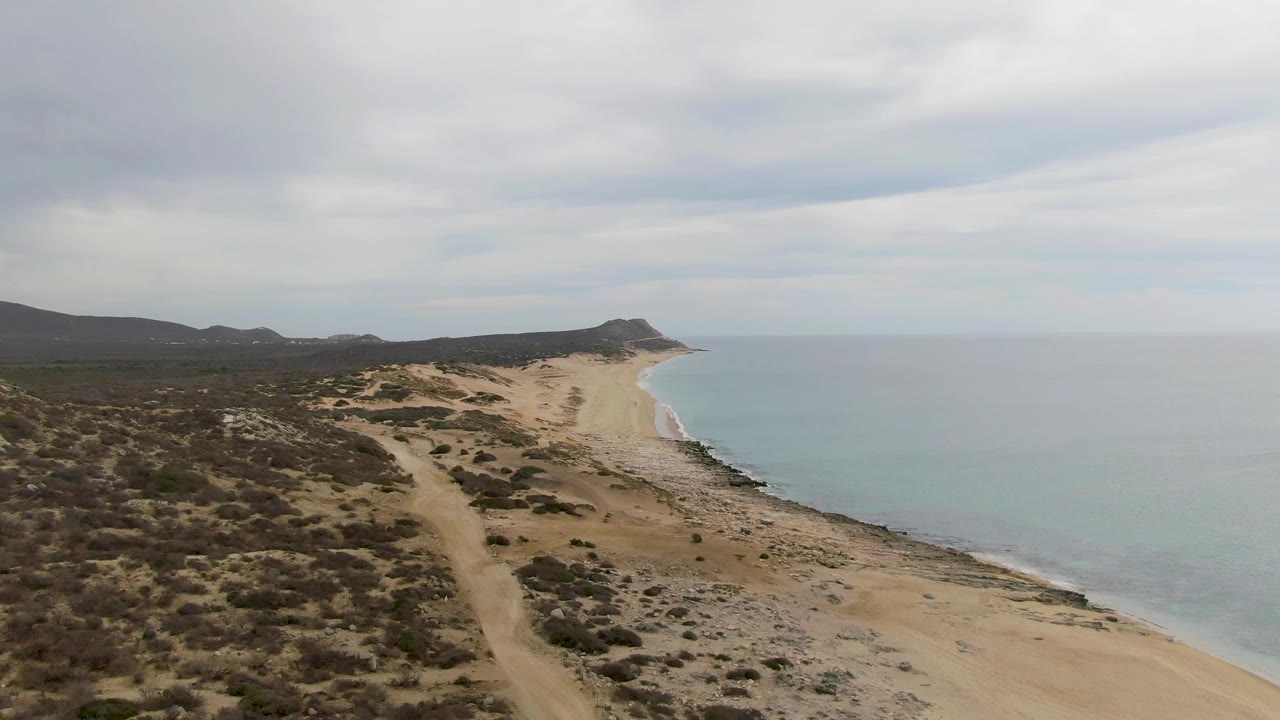 Elevated coastal view with dunes, dirt road, beach, calm ocean, and overcast clouds.