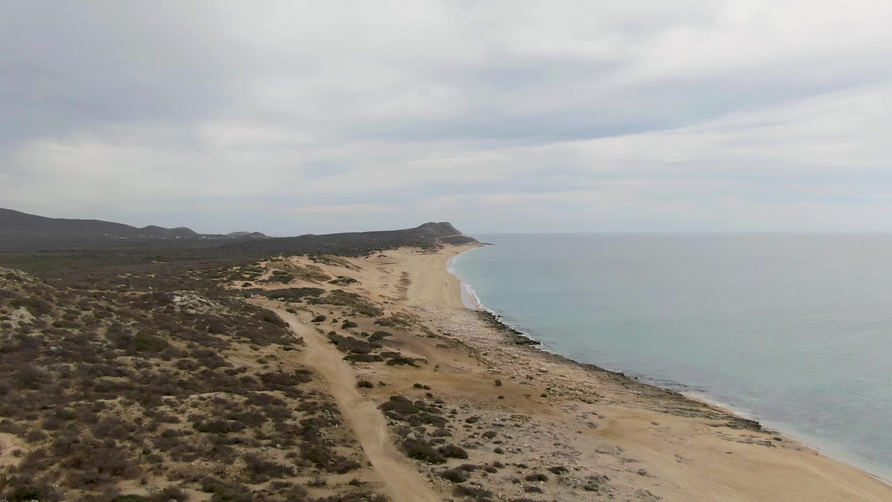 Aerial view along Punta Gorda Beach with scrub-covered dunes on the left, a pale sand strip curving along the water, distant headland shapes, and no visible people.