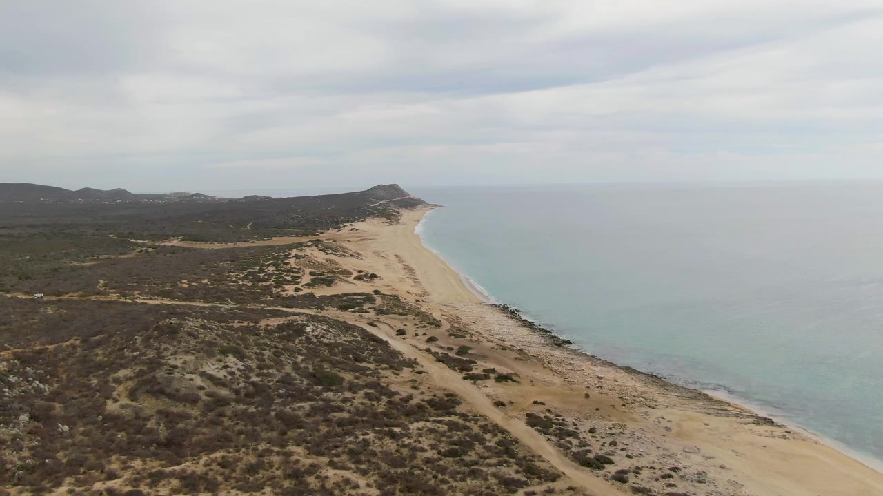 Opening aerial frame of Punta Gorda Beach in Baja California Sur, with dry coastal scrub in the foreground, a sandy beach curving beside calm ocean water, low hills in the distance, and no visible people.