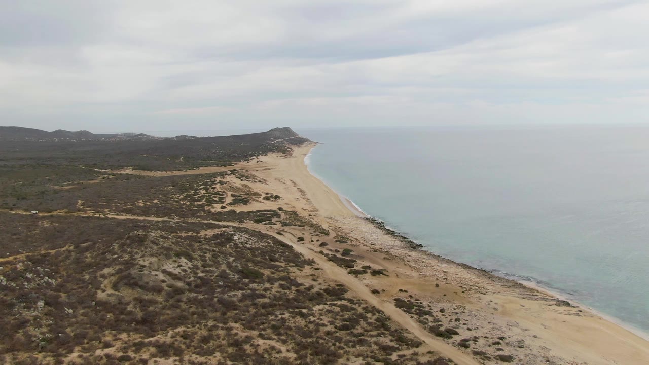 Second aerial frame over Punta Gorda Beach, showing more of the scrub-covered coastal terrain before the sandy shore, calm blue-green ocean to the right, distant low hills, and no visible people.