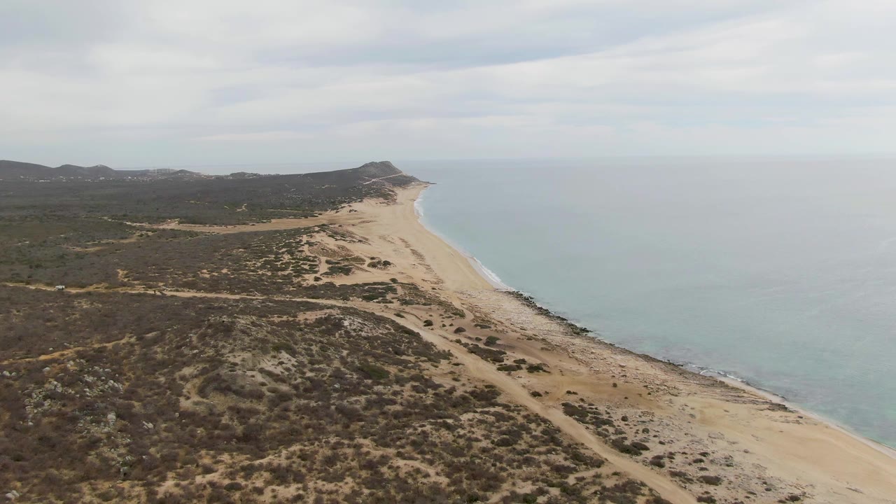 Coastal aerial view with sandy beach, pale water, dry brush, and distant hills