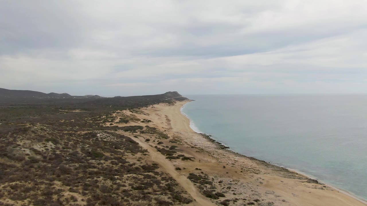 Early aerial frame over Punta Gorda Beach in Baja California Sur, showing scrub-covered dunes, a sandy shoreline, pale blue ocean, distant low hills, and no visible people.