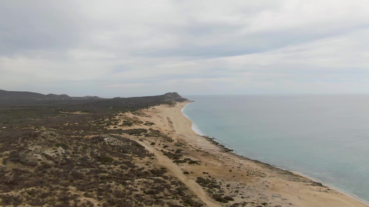 Sandy coastline with scrubby dunes, calm ocean, distant hills, and cloud cover.
