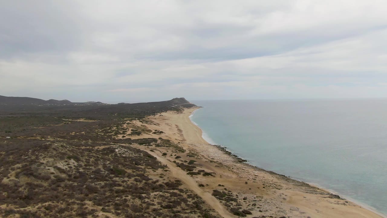 Mid-early frame from Punta Gorda Beach showing tan dune terrain in front, a narrow sandy shore beside blue-green water, dark hills inland, and no visible people.