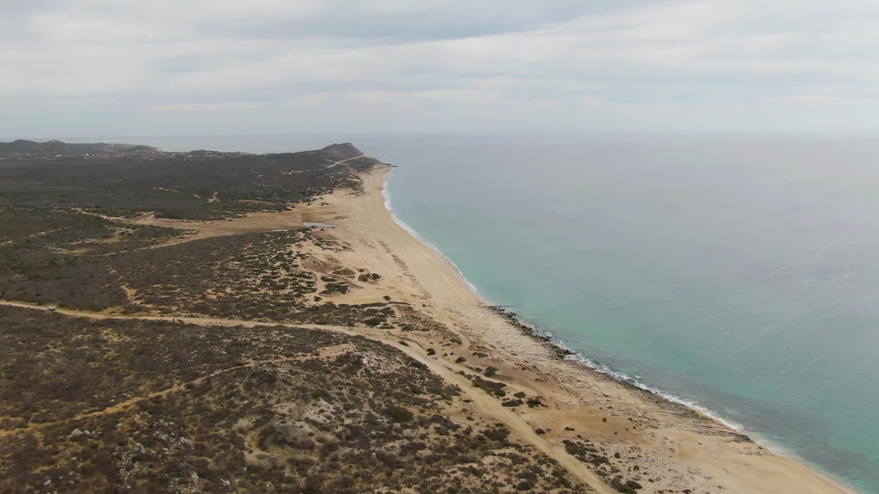 Aerial view along Punta Gorda Beach, showing a broad sandy strip between dark coastal vegetation and calm turquoise ocean, with distant hills under hazy cloud cover and no visible people.