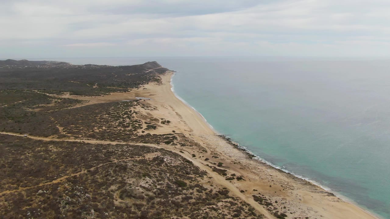 High drone frame over Punta Gorda Beach, with the beach running toward a low headland, scrubby dunes and dirt tracks inland, blue-green water offshore, and no visible people.