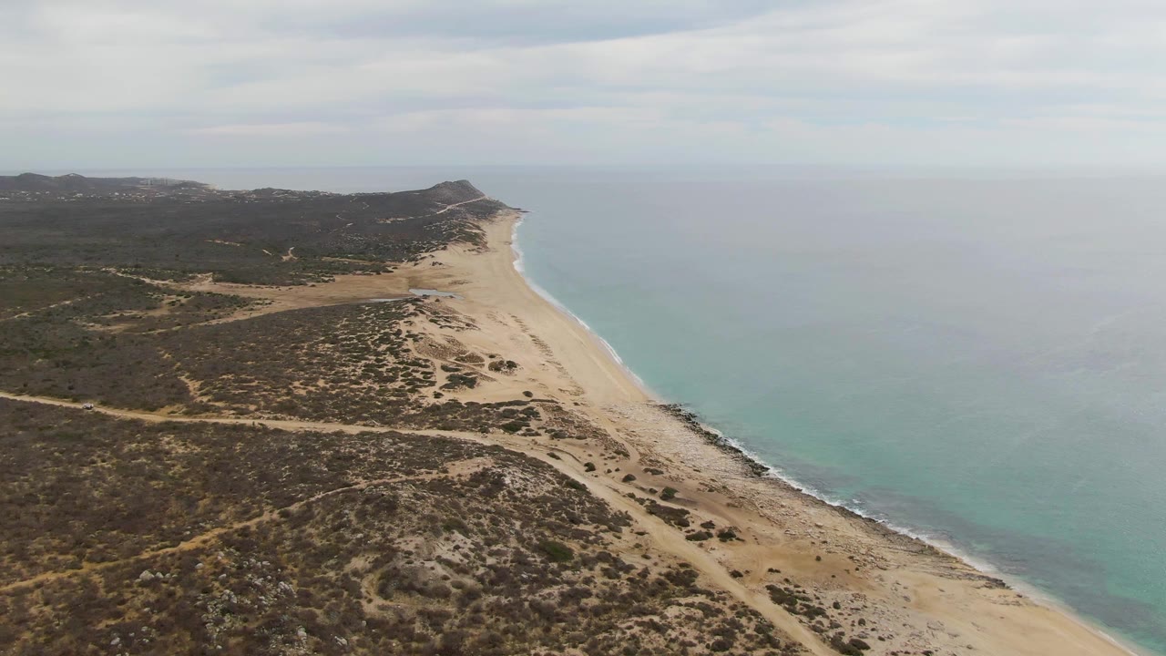 Aerial frame of Punta Gorda Beach with tan dunes and brush in the foreground, a thin surf line along the sand, turquoise sea to the right, distant hills, and no visible people.