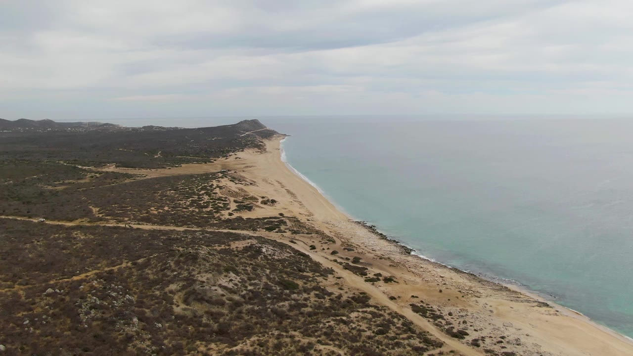 Elevated view over Punta Gorda Beach in Baja California Sur, with scrub-covered dunes in the foreground, a long sandy shoreline, turquoise ocean water, distant hills, and no visible people.