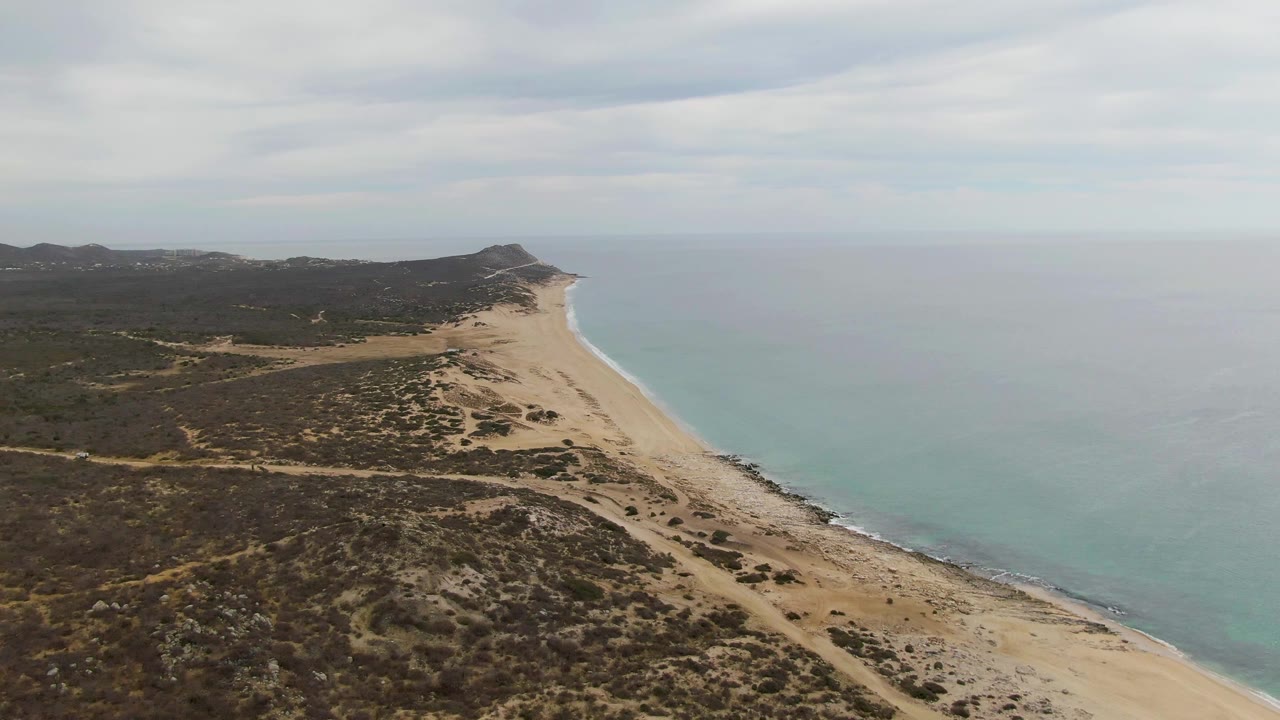 Aerial coastline view with beach, ocean, scrub terrain, and distant headland.